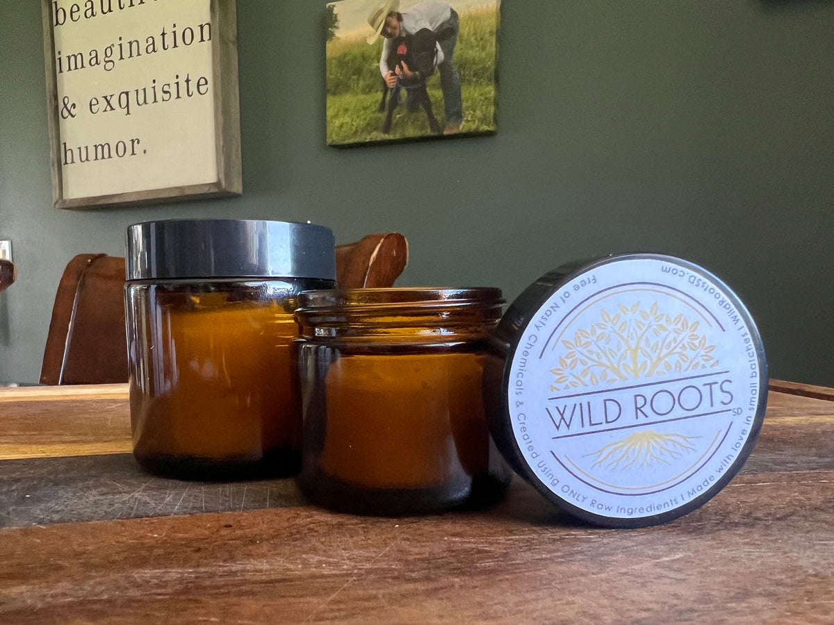 Two amber glass jar candles on a wooden table, with one jar open and its lid placed beside it. The lid label reads 'WILD ROOTS' with a tree logo.